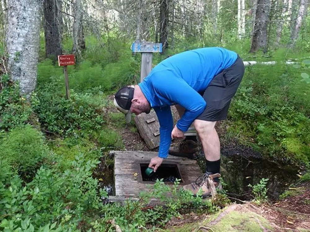 Man i blå tröja böjer sig ner för att skopa upp vatten ur en kallkälla i skogen
