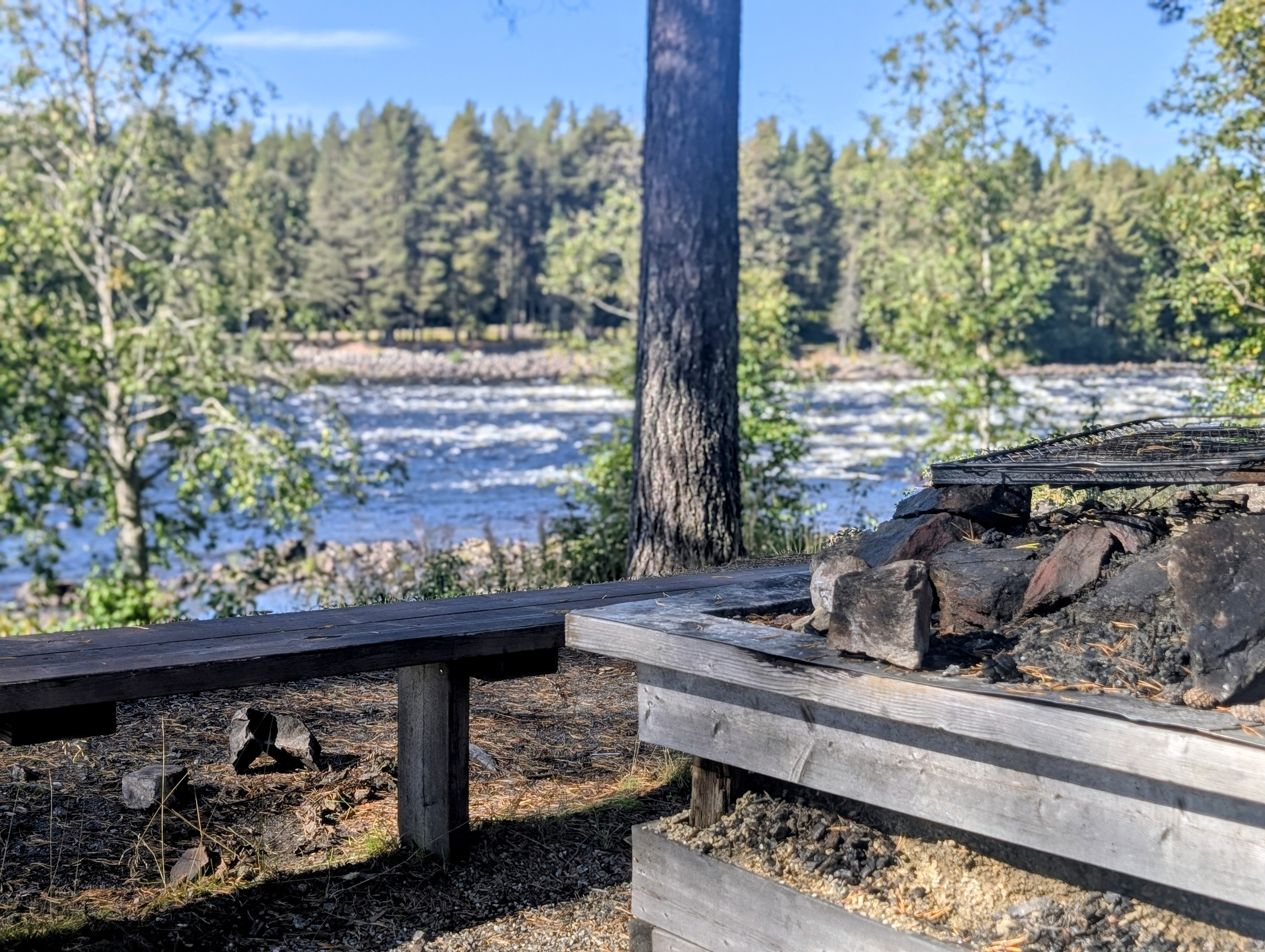 Grillplats med träbänkar intill Vindelälven och forsande vatten i bakgrunden.