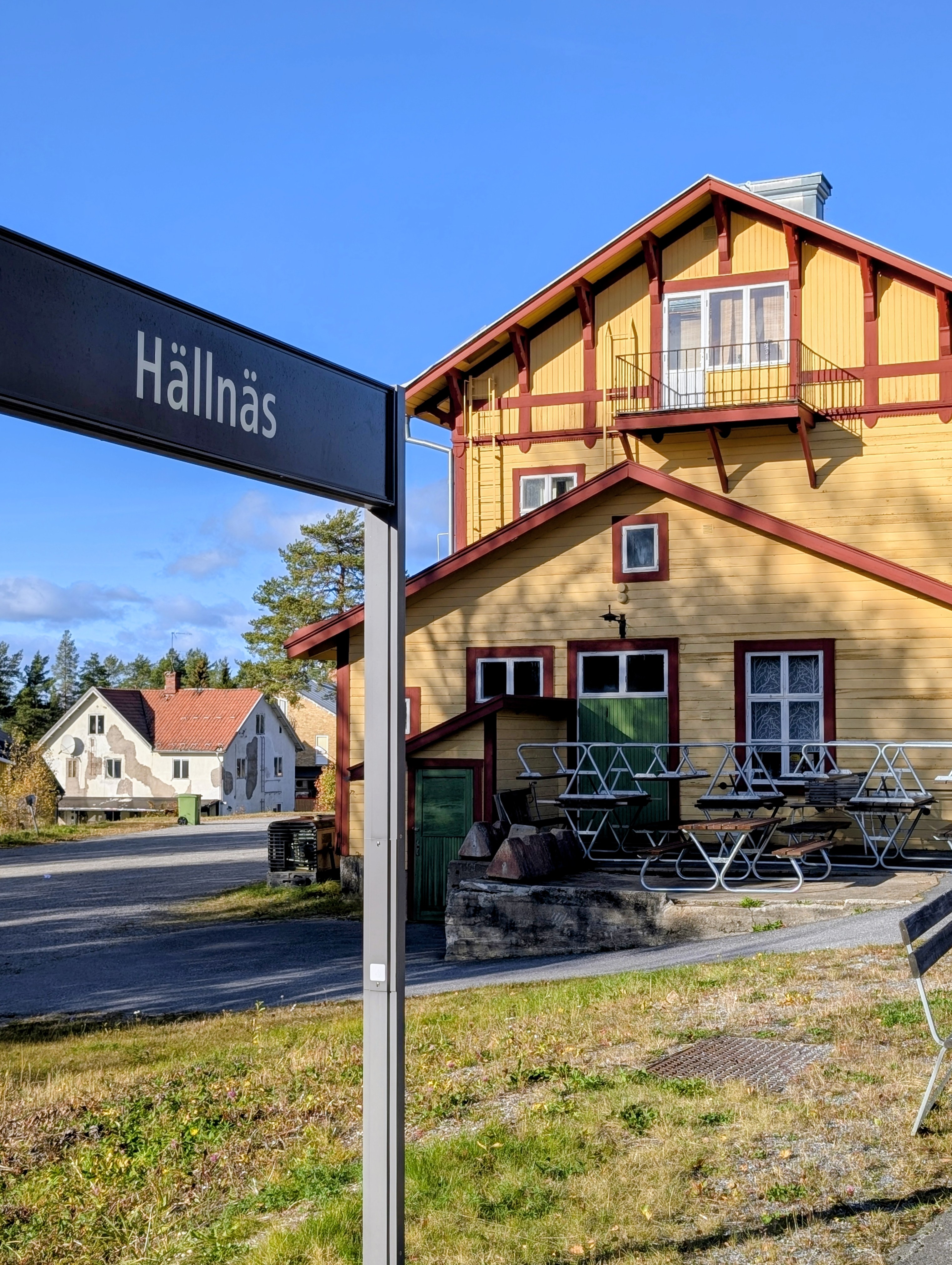 Stationsskylten Hällnäs framför det gulmålade stationshuset med röda detaljer.